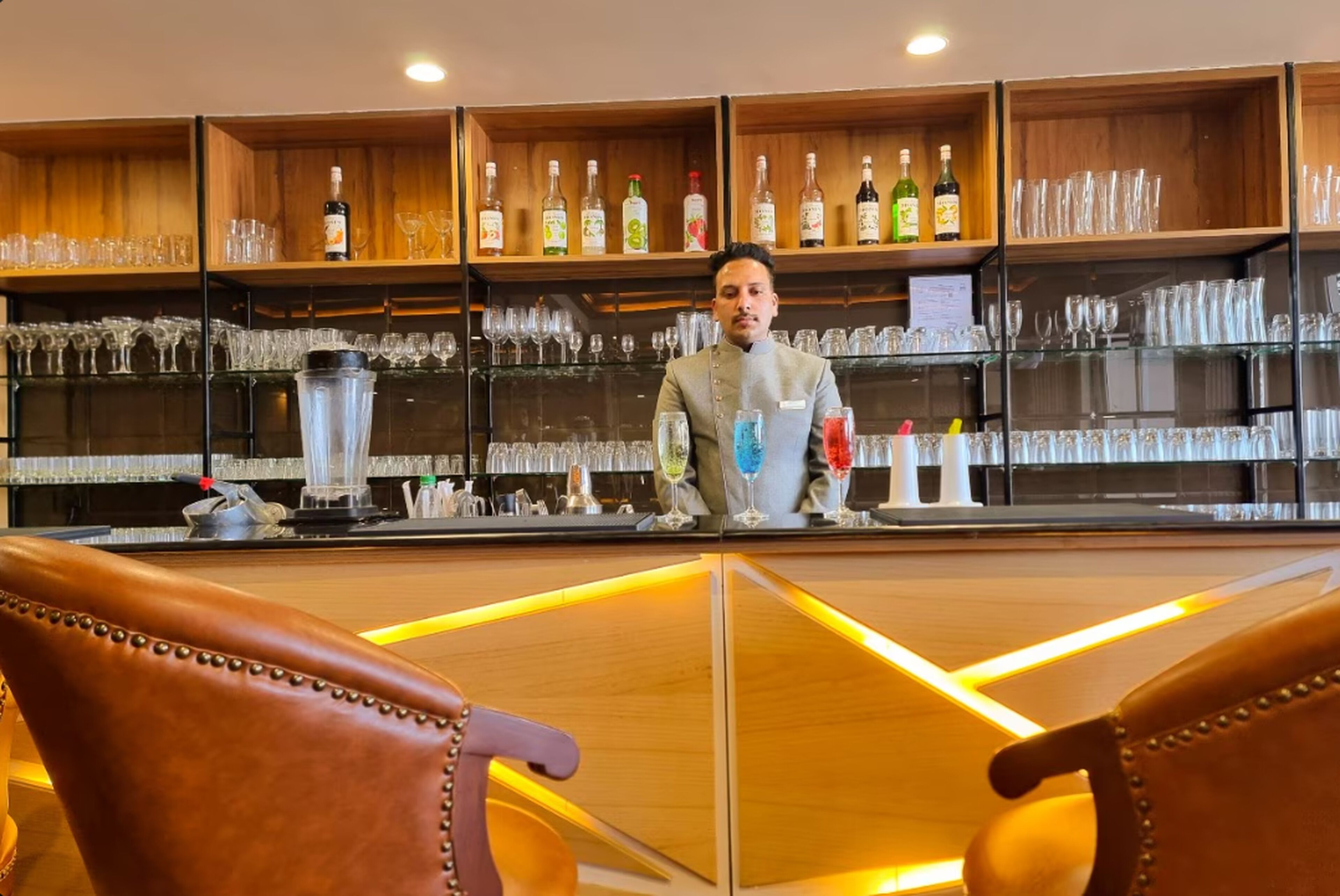 A bar area with shelves of bottles and glasses, a counter with a blender, and leather chairs.