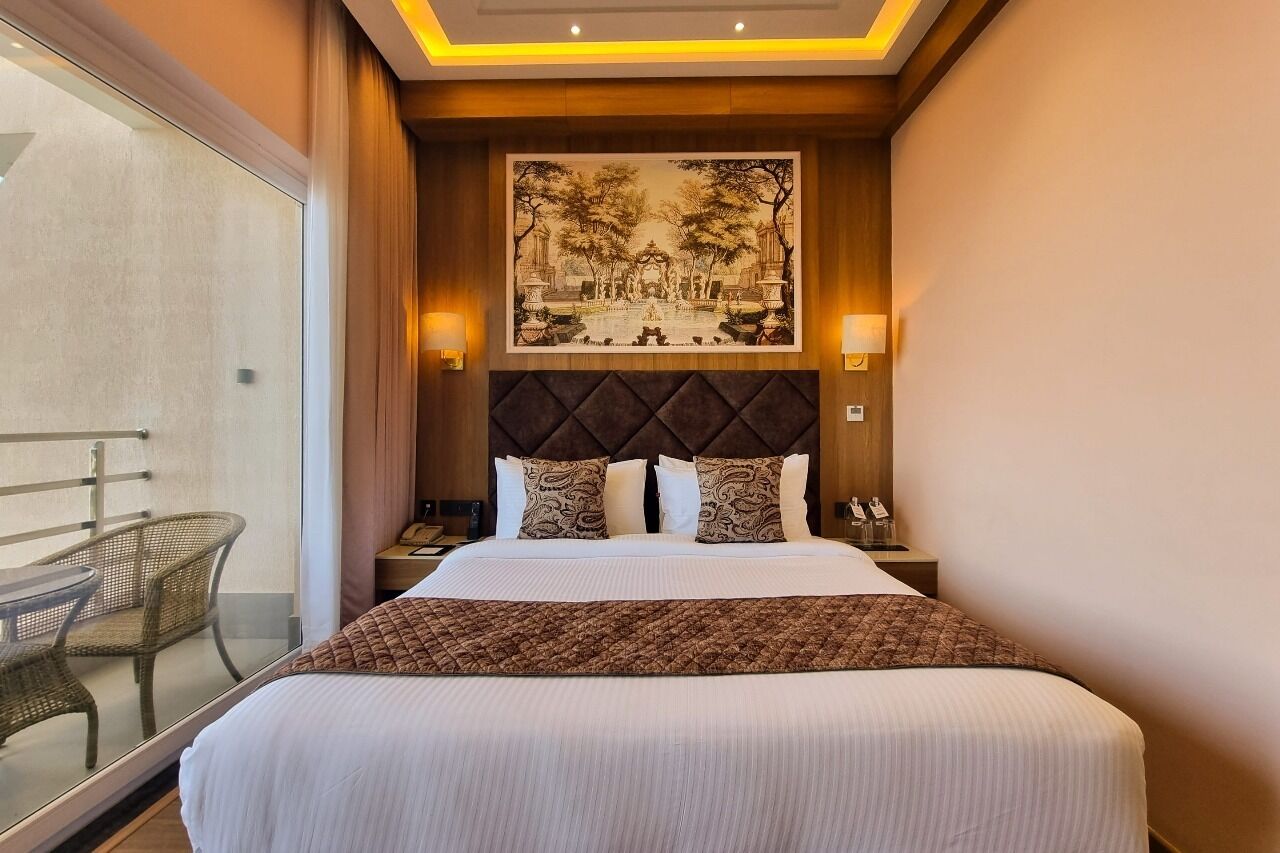 Habitación de hotel con una cama grande, un cabecero con un cuadro, dos lámparas de noche y vistas a un balcón con sillas de mimbre.