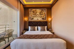 Habitación de hotel con una cama grande, un cabecero con un cuadro, dos lámparas de noche y vistas a un balcón con sillas de mimbre.