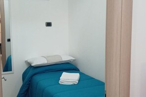 Ein kleines Zimmer mit einem Einzelbett, einer blauen Bettdecke, einem weißen Kopfkissen und einem gefalteten weißen Handtuch auf dem Bett.