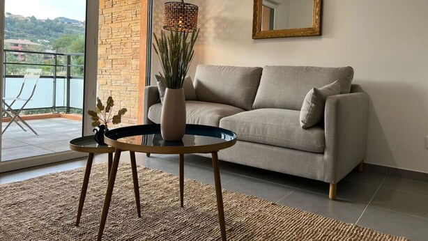Una sala de estar con sofá, una mesa redonda con un jarrón, un espejo grande y vistas a un balcón con una silla.
