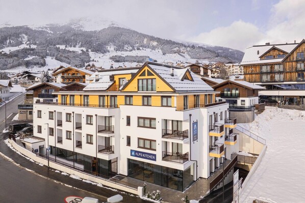 Un hotel de varios pisos con balcones, un letrero que dice 'ALPS KITCHEN' y una montaña nevada al fondo.