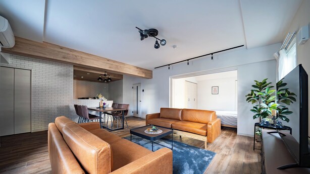 A modern living room with a leather sofa, a coffee table, and a TV. There is a dining area with a table and chairs, and a kitchen in the background.