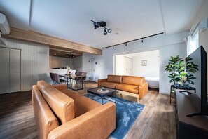 A modern living room with a leather sofa, a coffee table, and a TV. There is a dining area with a table and chairs, and a kitchen in the background.