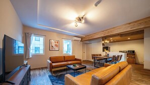 A modern living room with a sofa, coffee table, and TV. It includes a dining area with chairs and a kitchen in the background.