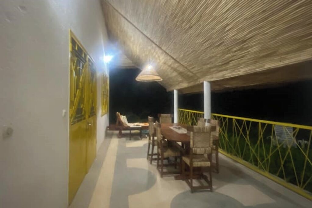 Un área de comedor al aire libre con mesas y sillas, techo de paja y barandilla amarilla.