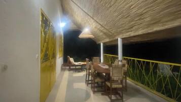 Un área de comedor al aire libre con mesas y sillas, techo de paja y barandilla amarilla.