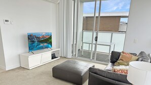 A modern living room with a flat-screen TV on a white entertainment unit, a grey sofa, a black ottoman, and a wicker side table. - Bright Zetland Apartment with CBD Views and Fast Free WiFi (Zetland)