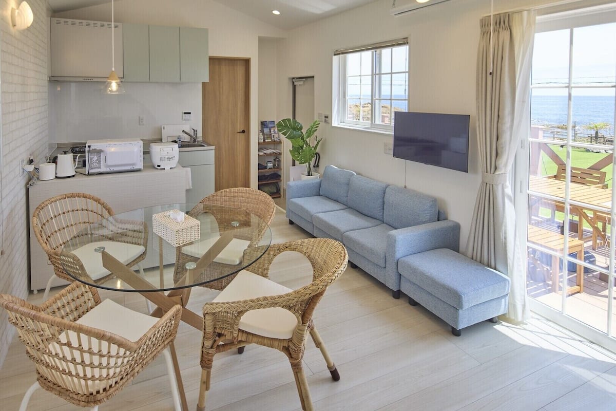 A modern living room with a glass dining table, wicker chairs, a blue sofa, and a TV mounted on the wall.