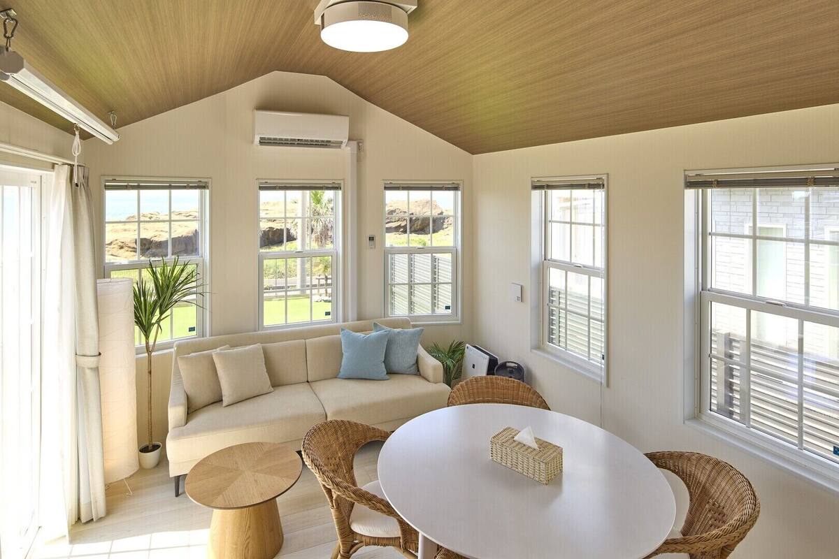 A spacious living room with a high ceiling, large windows, a beige sofa, a round table, and wicker chairs.