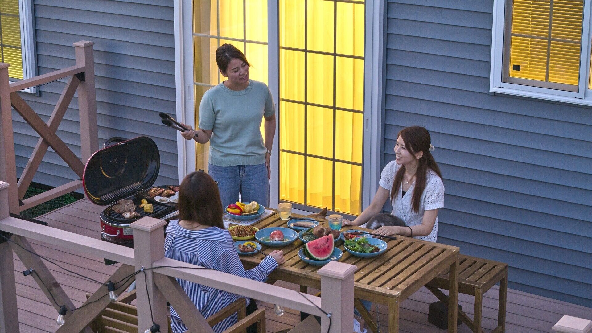 An outdoor dining area with a grill, wooden deck, and a house with blue siding and yellow windows.