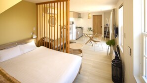 A bedroom with a large bed, a wooden partition, a dining area with chairs, a kitchen in the background, and a window with curtains. - Solana Shirahama (Minamiboso)