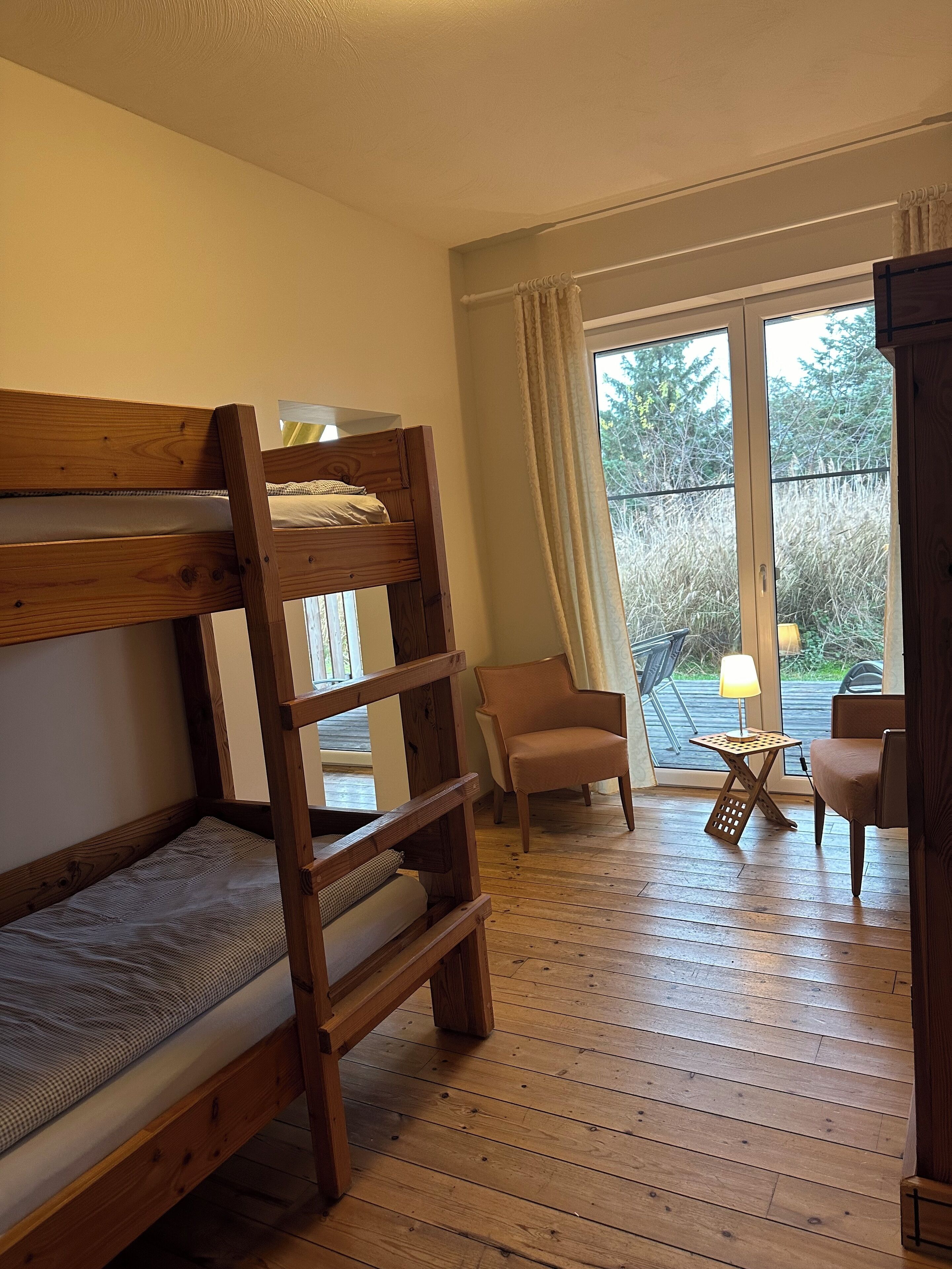 A room with a bunk bed, a chair, a small table with a lamp, and a view through a glass door.