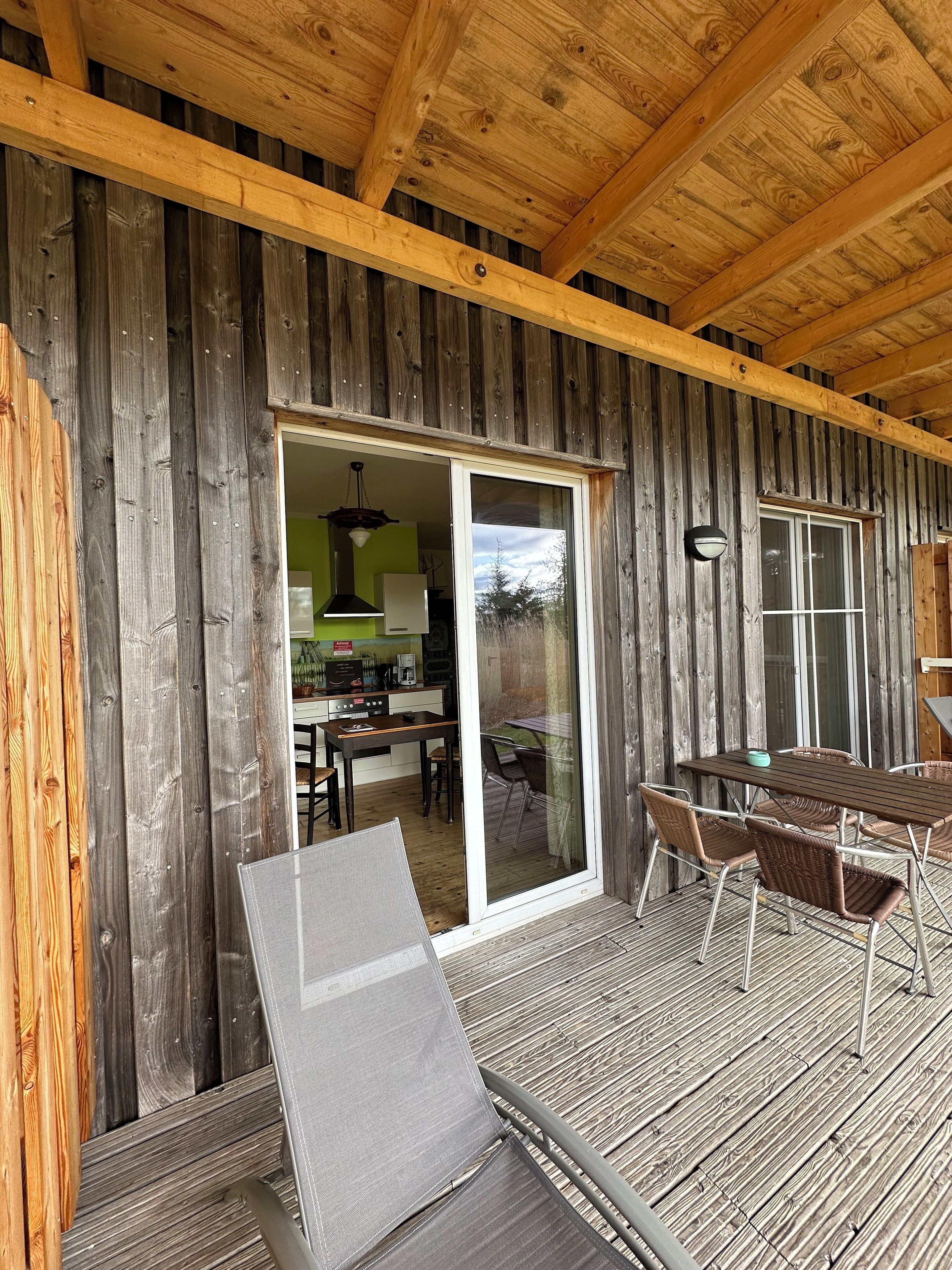 A wooden deck with a dining area, a sliding glass door leading to a kitchen, and a ceiling with exposed beams.