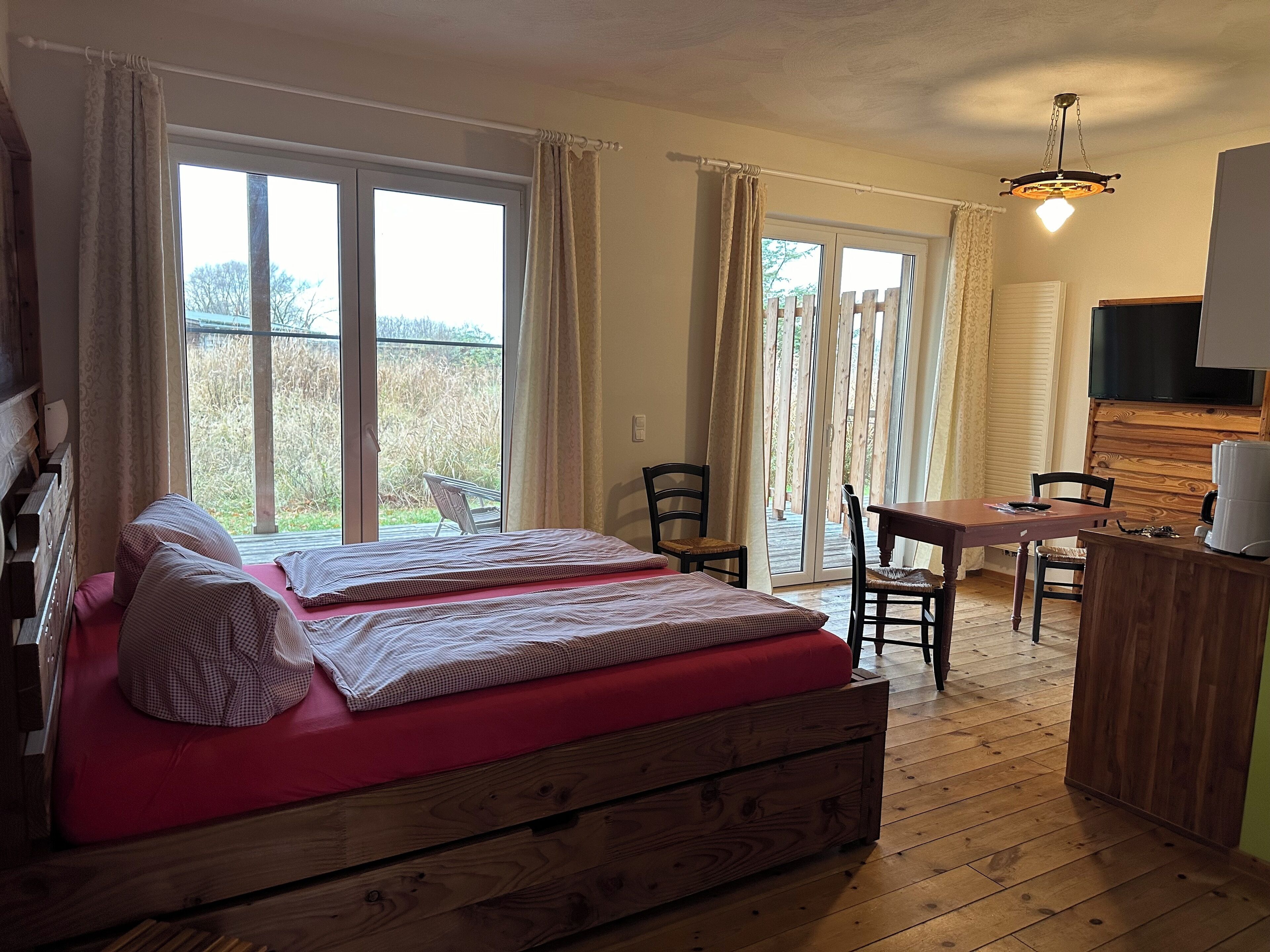 A bedroom with two beds, a wooden desk, a chair, a television, and a balcony with curtains.