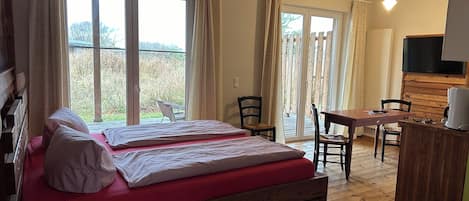 A bedroom with two beds, a wooden desk, a chair, a television, and a balcony with curtains.