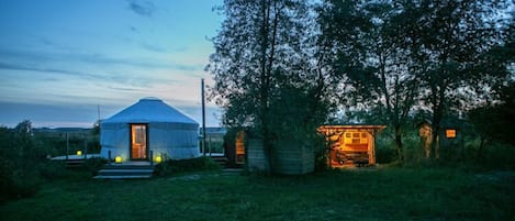 A yurt with a door and steps leading up to it, surrounded by trees and a small wooden structure with a lit interior.