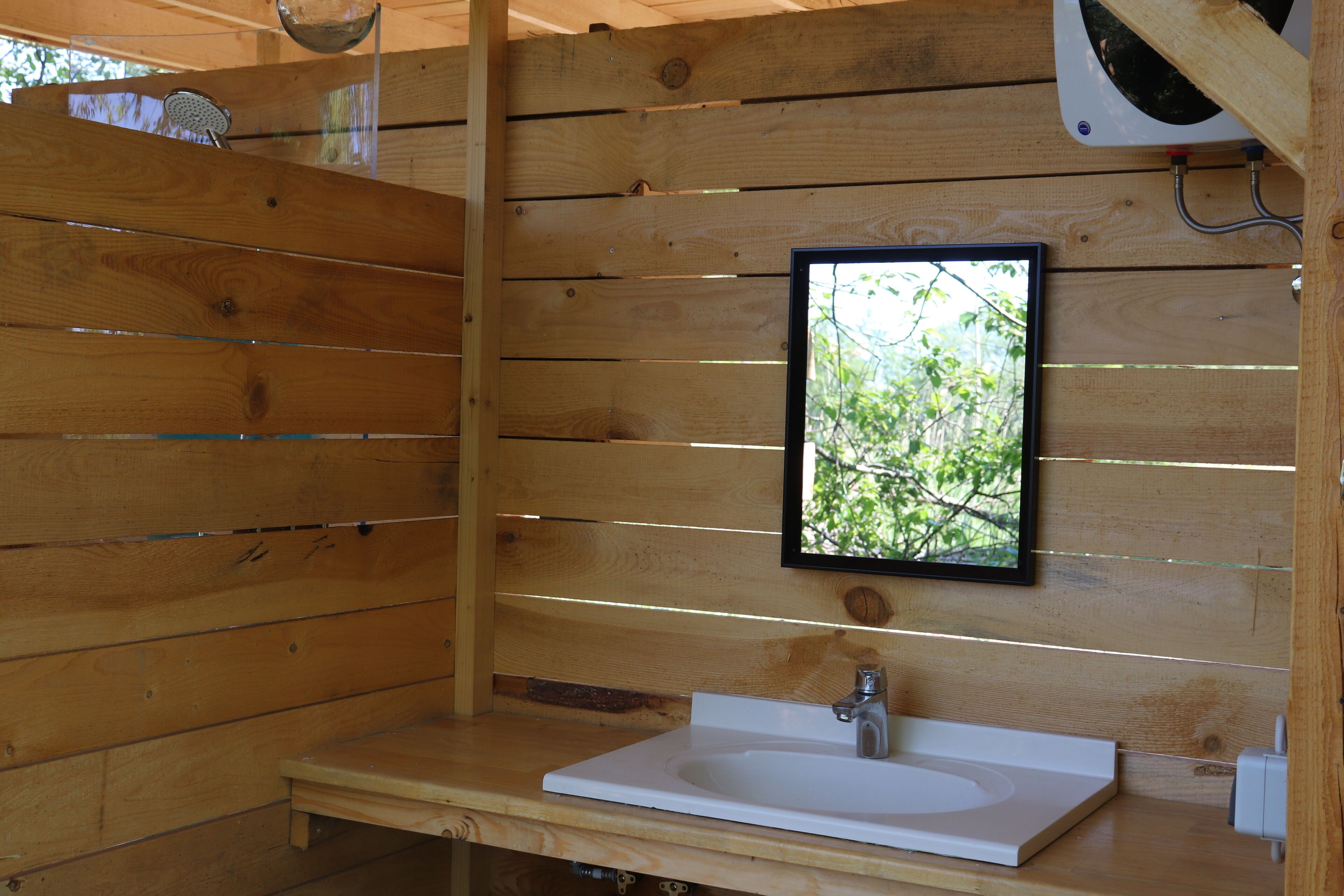 Una cabaña de madera con un lavamanos blanco, una ventana con vistas a árboles y un área de ducha.