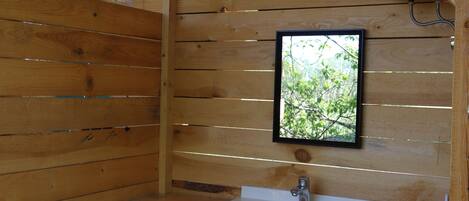 Una cabaña de madera con un lavamanos blanco, una ventana con vistas a árboles y un área de ducha.