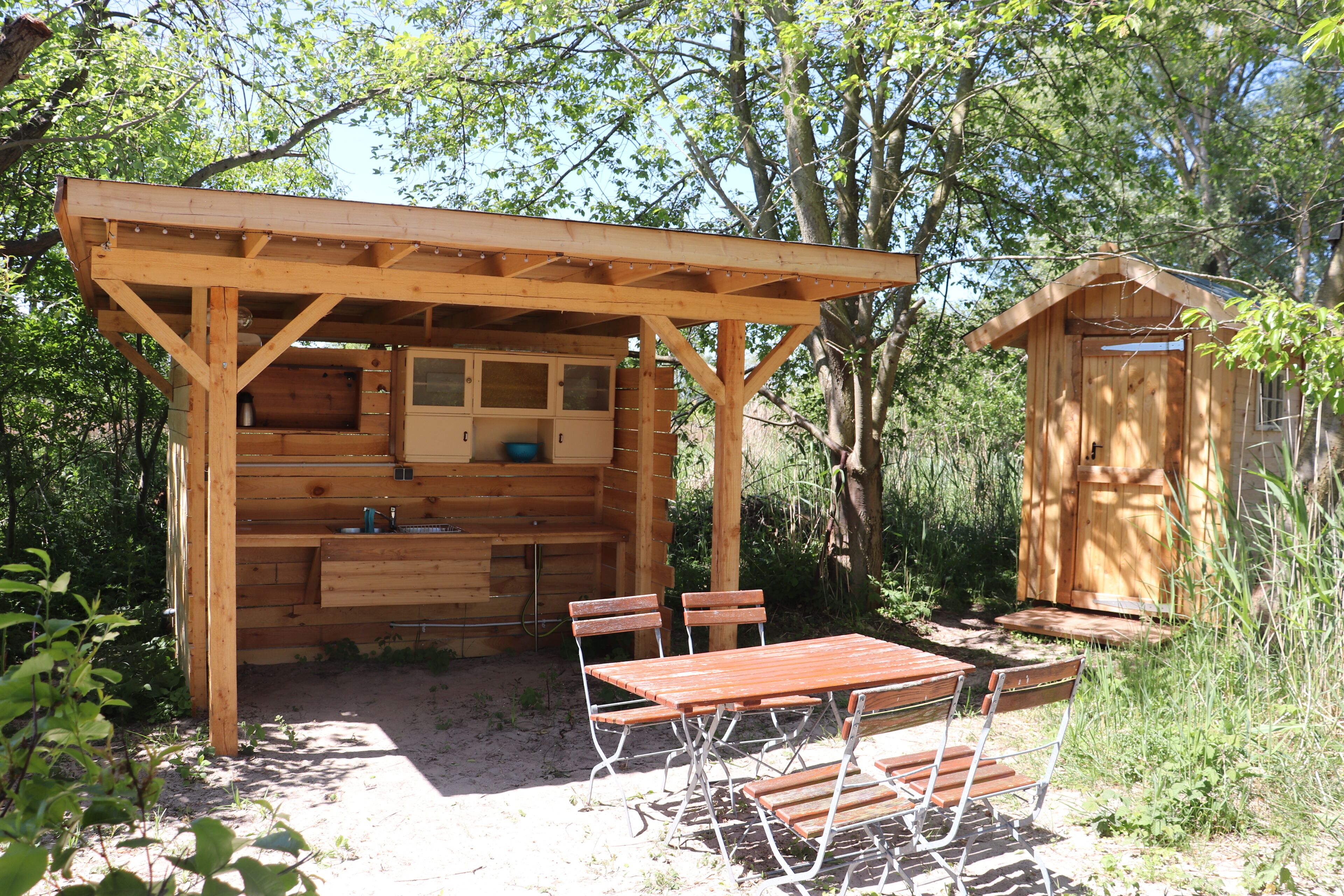 Une cuisine extérieure en bois, avec une table et des chaises, entourée d’arbres.