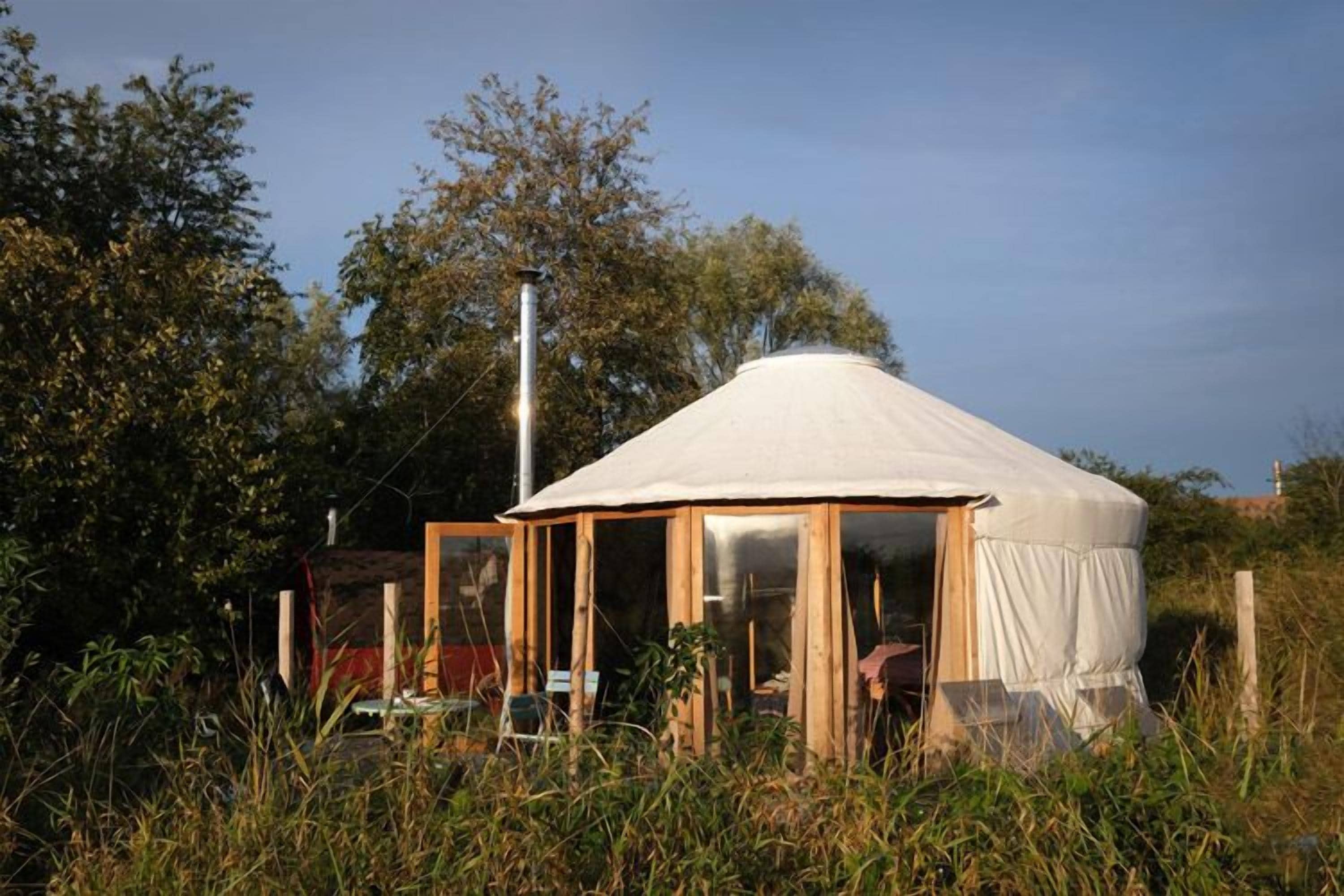 Una yurta con techo cónico blanco, estructura de madera y puertas de vidrio, rodeada de hierba alta y árboles.