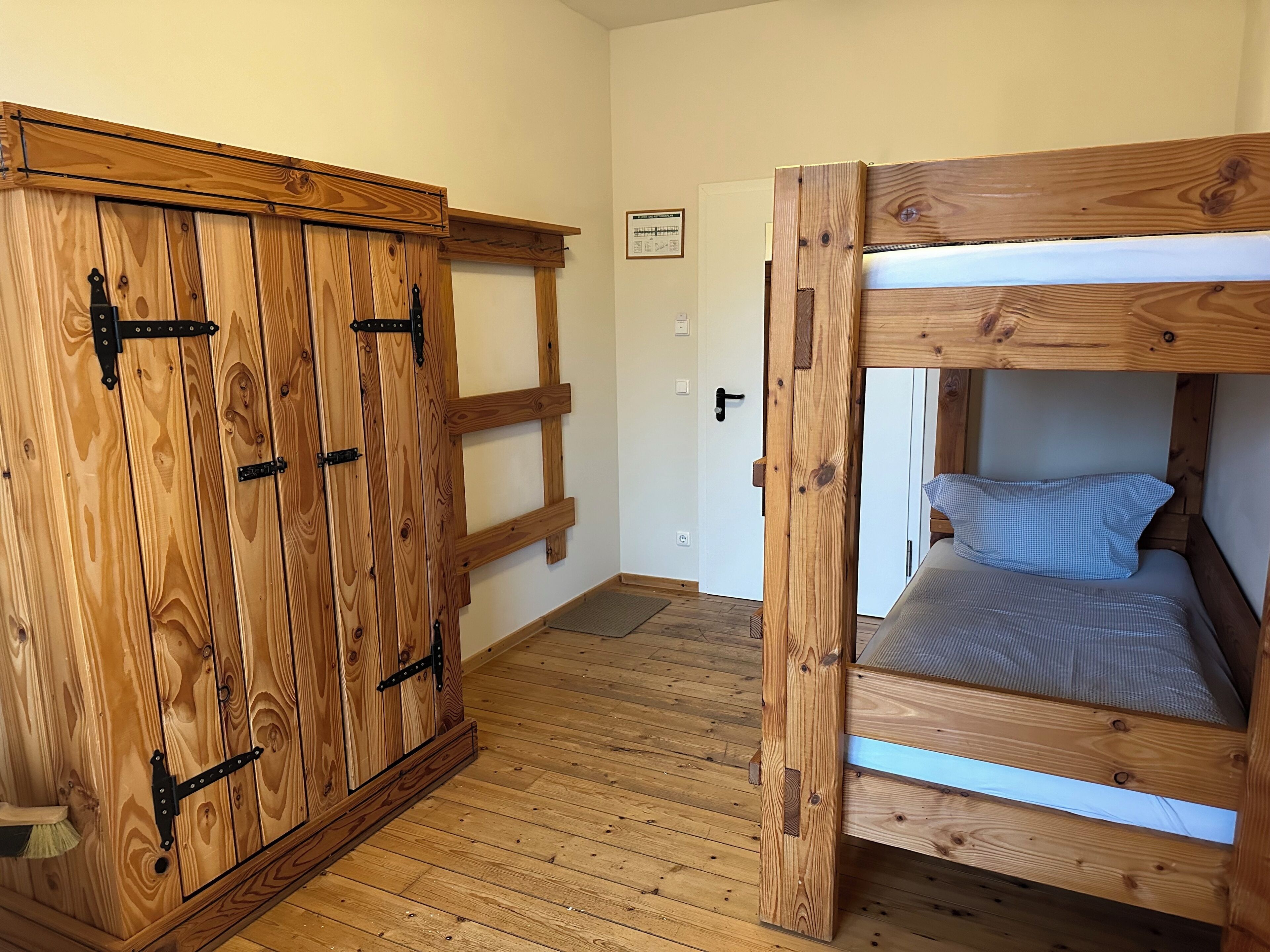 A wooden bunk bed with a mattress and pillows, a wooden wardrobe with multiple doors, and a door leading to another room.