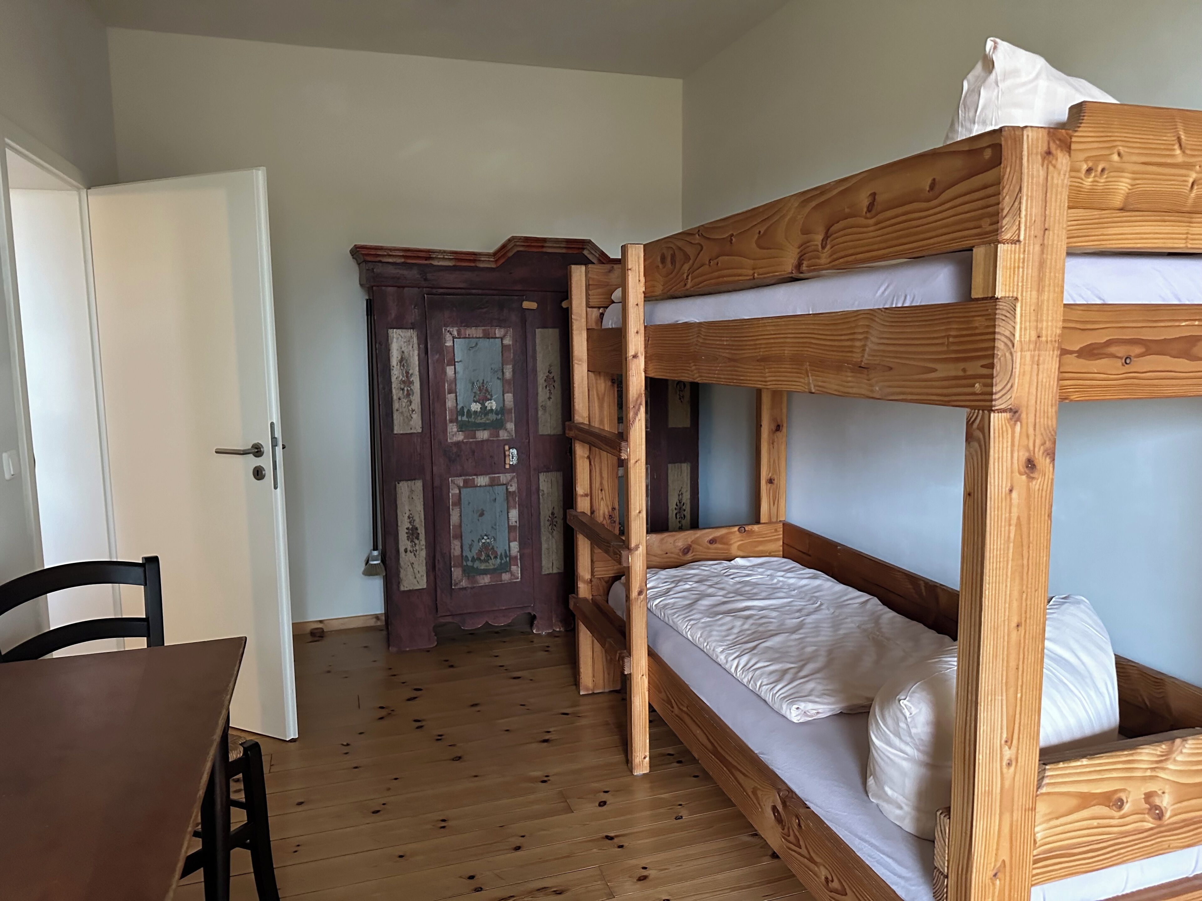 A bunk bed with a wooden frame and white bedding, a wooden table with a chair, and a door with floral decorations.