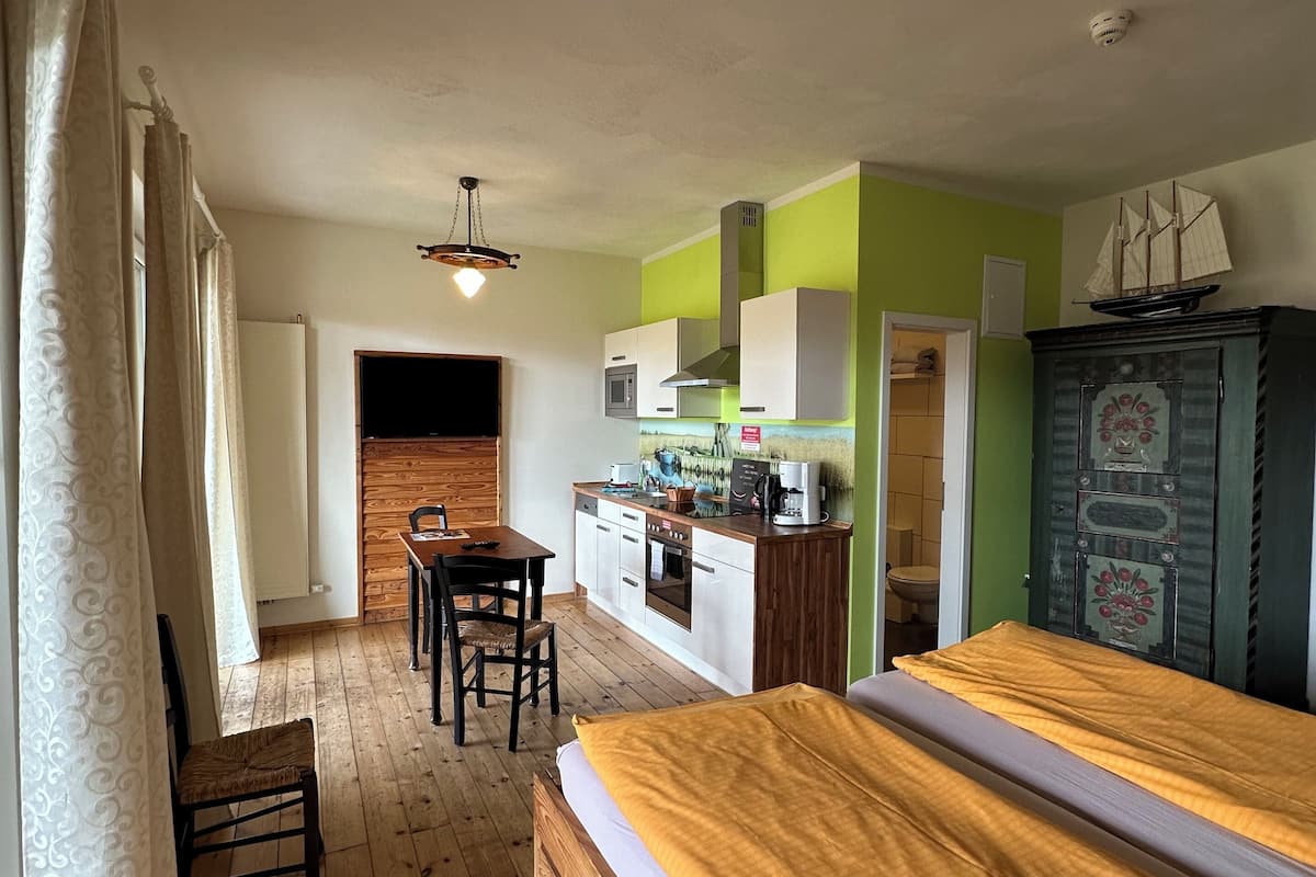 A room with a bed, a small table with a chair, a kitchenette, and a cabinet with a ship decoration.