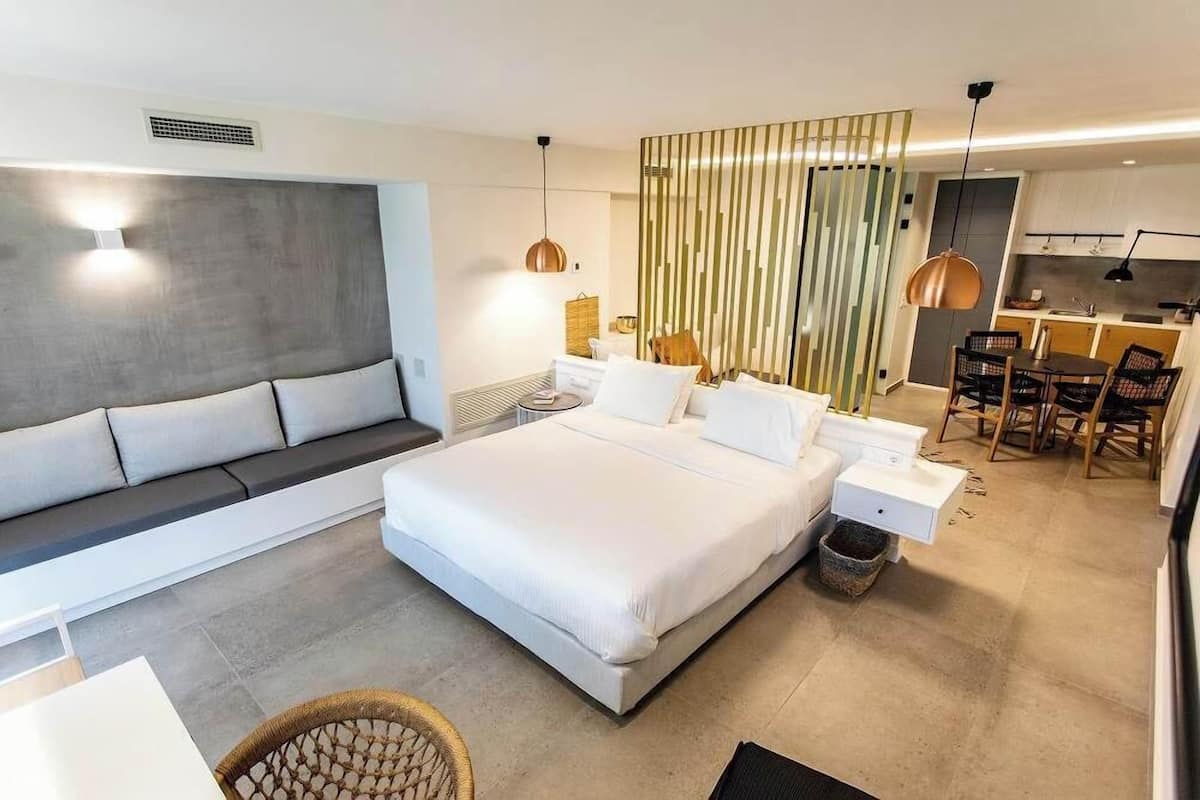 A modern hotel room with a large bed, a sofa, a dining area, and a kitchen.
