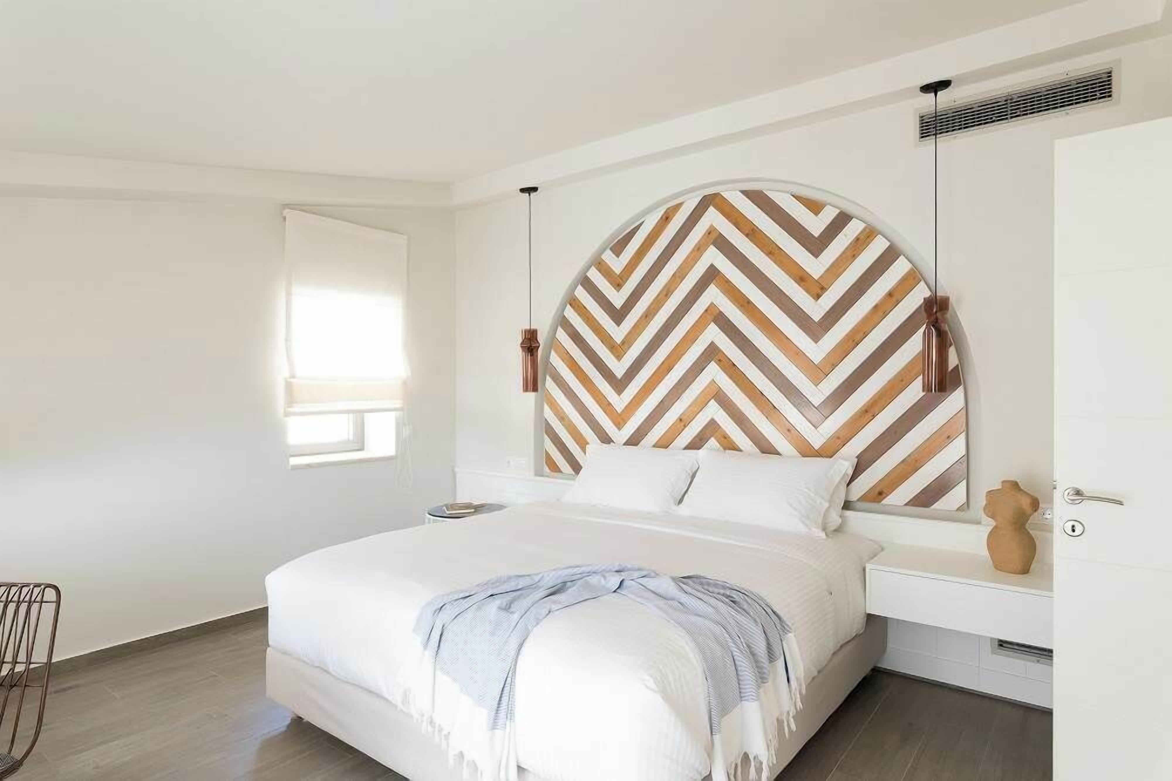 A modern bedroom with a large bed, a wooden chevron-patterned headboard, a bedside table, and a window with blinds.