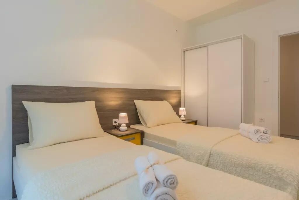 A hotel room with two beds, each with a headboard, bedside tables with lamps, and rolled towels on the beds.