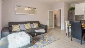 Un salon moderne avec un canapé gris, des coussins à motifs, une petite table d’appoint blanche et un pouf blanc moelleux. Un lustre est suspendu au plafond, et on aperçoit à l’arrière-plan un coin repas avec une table et des chaises.