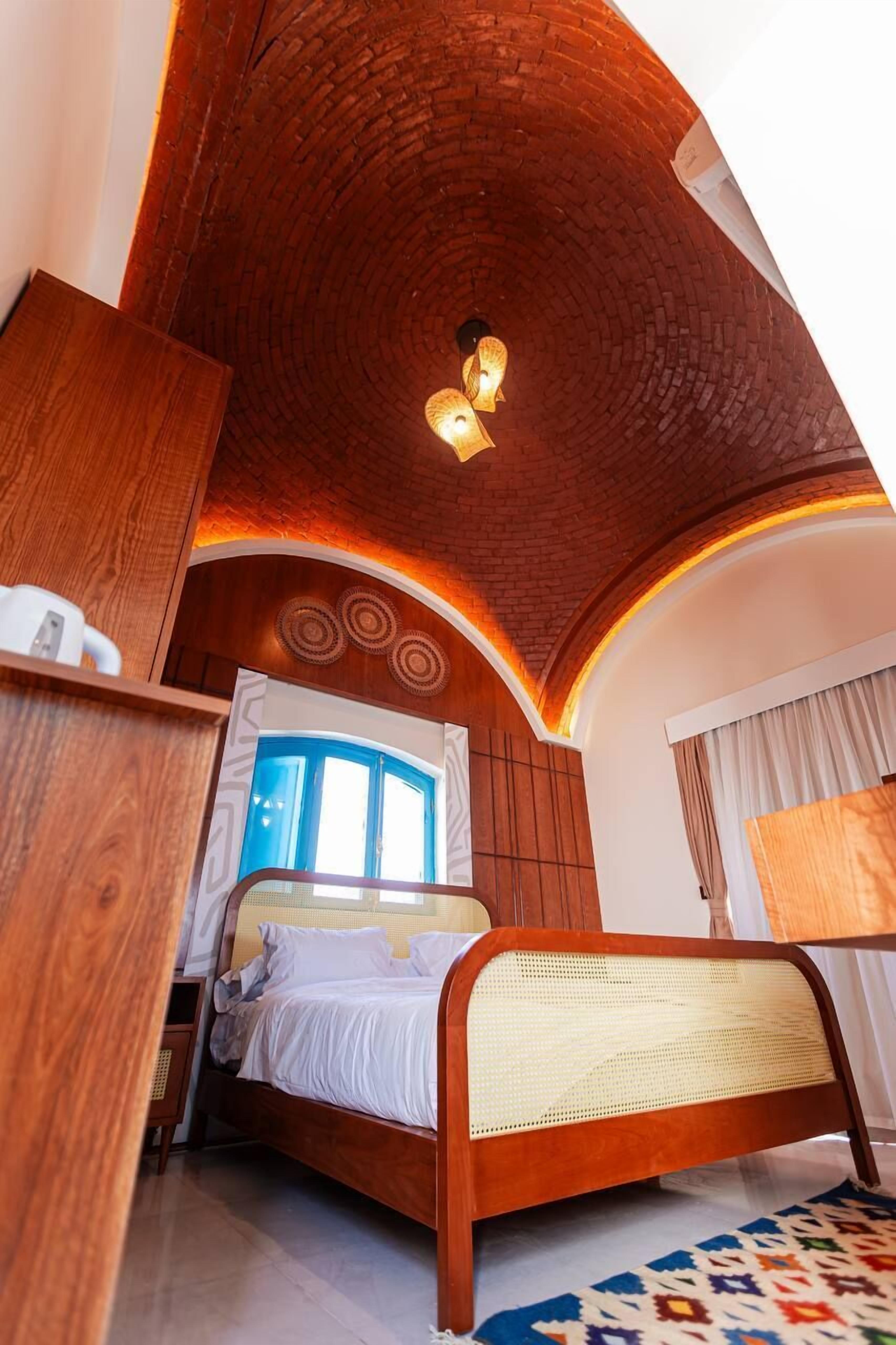 Ein Schlafzimmer mit einem hölzernen Kopfteil, einem Bett mit weißer Bettwäsche, einem Fenster mit blauen Vorhängen und einem gemusterten Teppich.