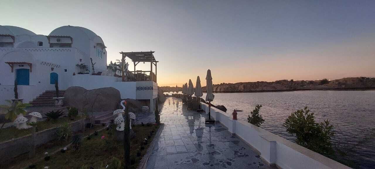 Propiedad frente al agua con edificios blancos, un área de estar cubierta al aire libre y un sendero pavimentado a lo largo del agua.