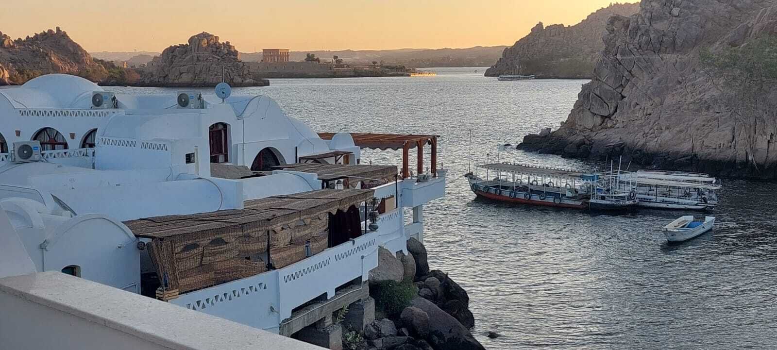 Un edificio frente al mar con balcón que da a una masa de agua, una embarcación fondeada cerca y acantilados rocosos al fondo.