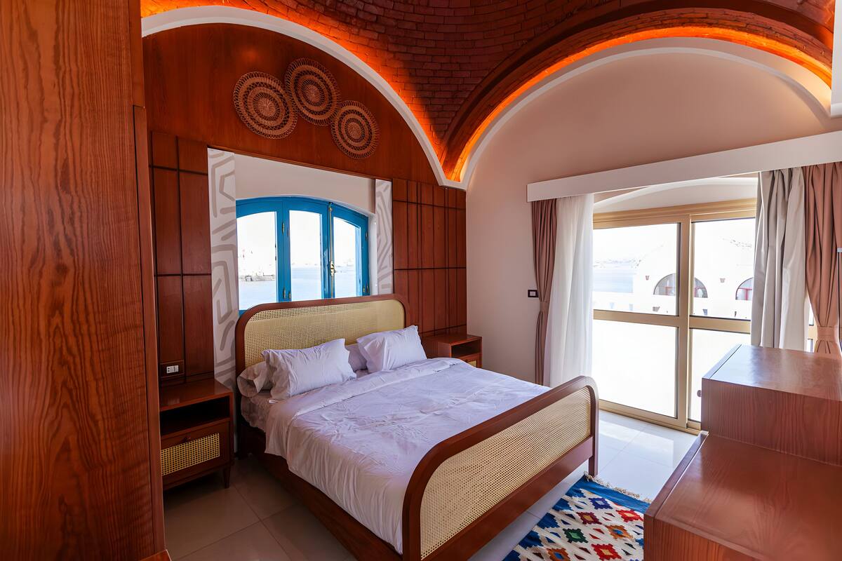 Un dormitorio con cabecera de madera, una cama grande, una mesita de noche, un aparador y vistas al exterior a través de una puerta corrediza de vidrio.