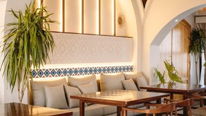 Ein gemütlicher Lounge-Bereich mit einer Couch, Holztischen und Stühlen. Übertöpfe mit Pflanzen und ein dekoratives Wandpaneel sind ebenfalls vorhanden.