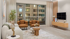 Un salón moderno con un sofá, sillones, una mesa de centro y un televisor. Cuenta con un balcón con barandilla y vistas a un edificio.