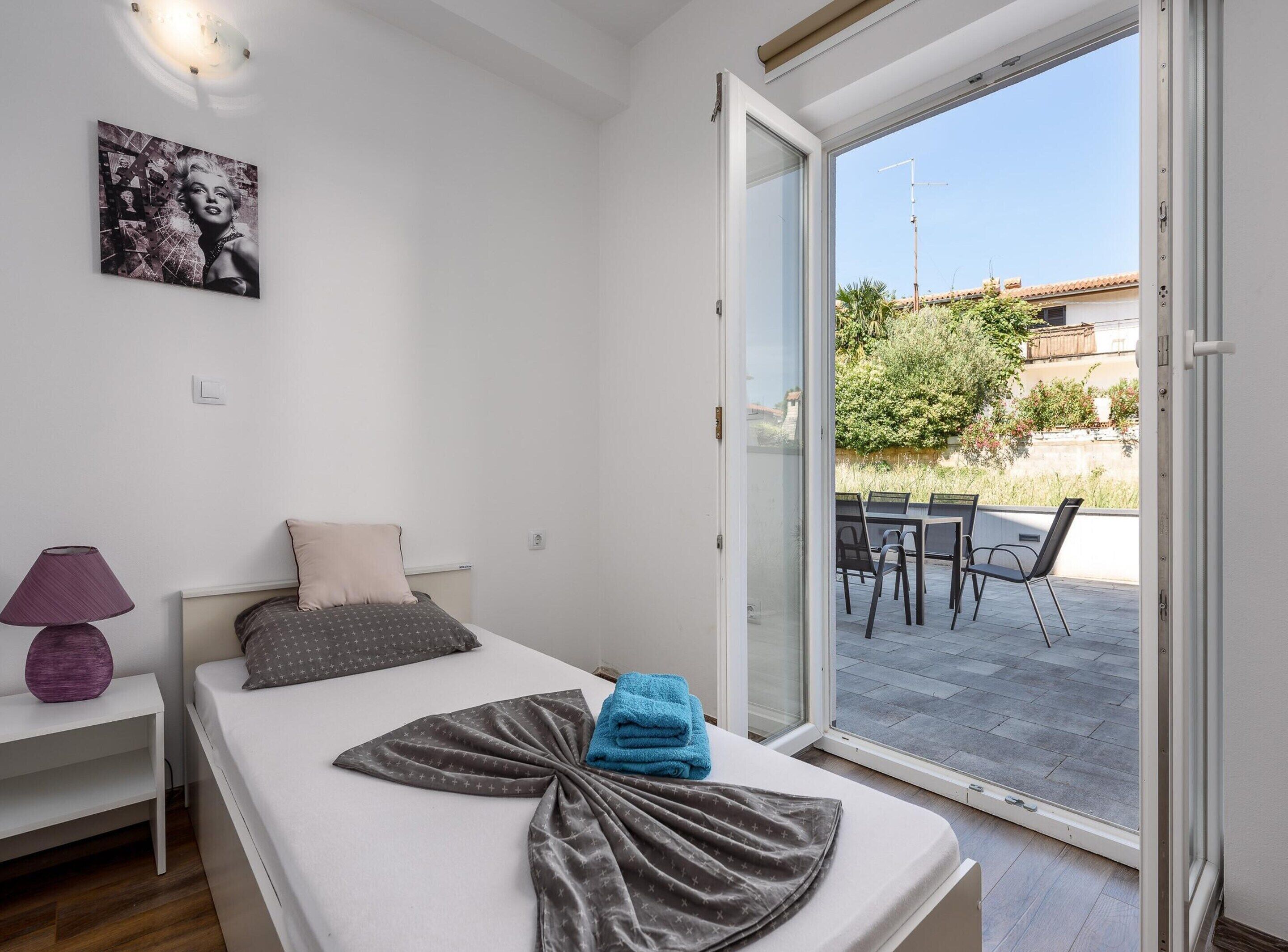 A bedroom with a bed, a nightstand, a lamp, a painting, and a sliding glass door to an outdoor patio.