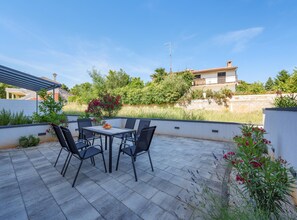 Eine Terrasse mit Tisch und Stühlen, umgeben von Pflanzen und Blick auf ein Gebäude.