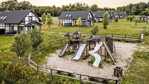 Een speeltuin met glijbanen en een houten omheining, omringd door huizen met donkere daken en witte muren.