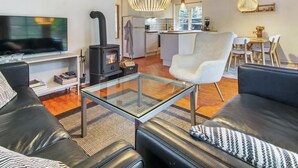 Ein modernes Wohnzimmer mit einem schwarzen Sofa, einem Glastisch und einem weißen Sessel. Im Hintergrund steht ein schwarzer Kaminofen, und es gibt eine Küchenzeile mit einem Esstisch und Stühlen.