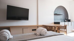 A modern hotel room with a flat-screen TV mounted on the wall, a wooden desk, a kitchen area with a sink, and a bed with a rolled-up towel. - Alinea Lano Old Town Center (Limassol)