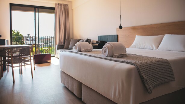 A hotel room with a large bed, a desk, and a chair. There is a balcony with a view of the city. - Alinea Lano Old Town Center (Limassol)