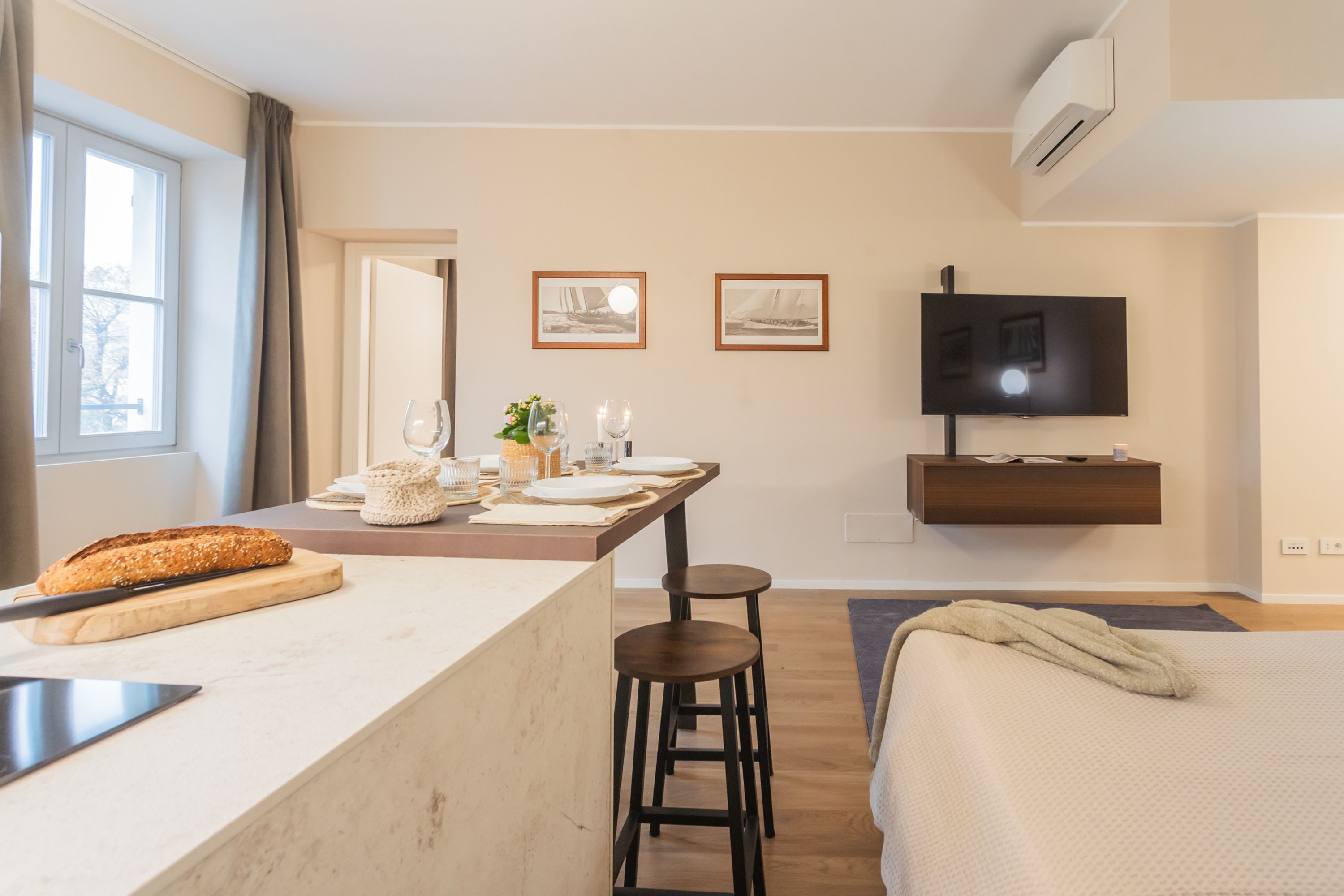 Une chambre d’hôtel moderne dotée d’un coin cuisine, d’une table à manger dressée avec des assiettes et des verres, d’un lit avec une couverture et d’un téléviseur fixé au mur.
