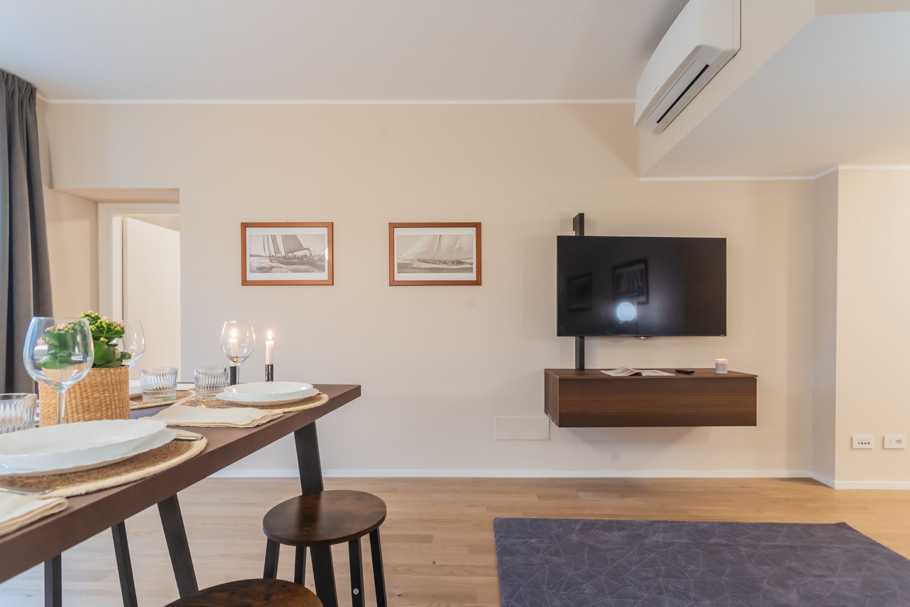 A modern living room with a dining area, a flat-screen TV mounted on the wall, and a wooden dining table set with plates, glasses, and candles.
