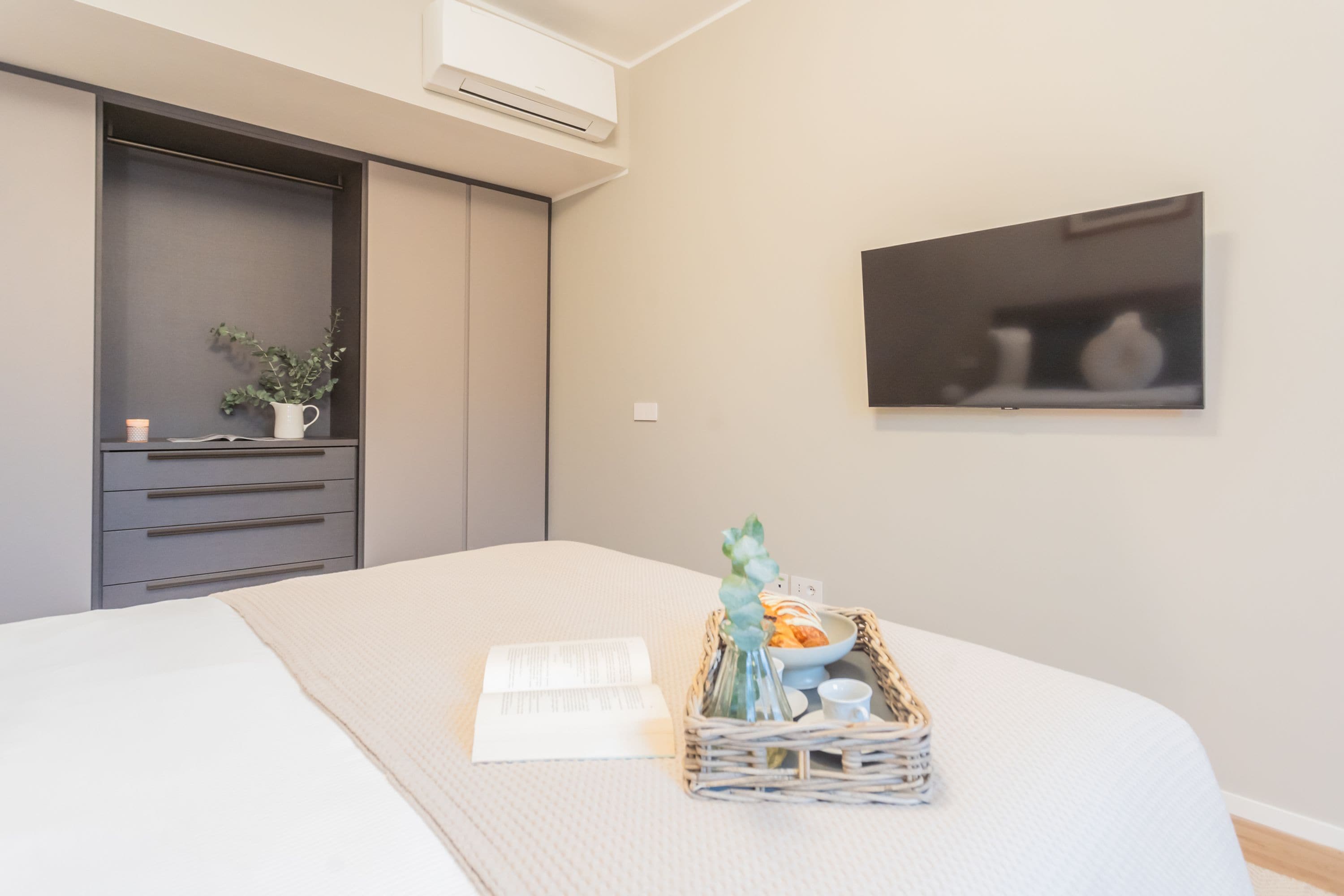 A modern bedroom with a bed, a bedside table with a book and a vase, a wall-mounted TV, and a wardrobe.