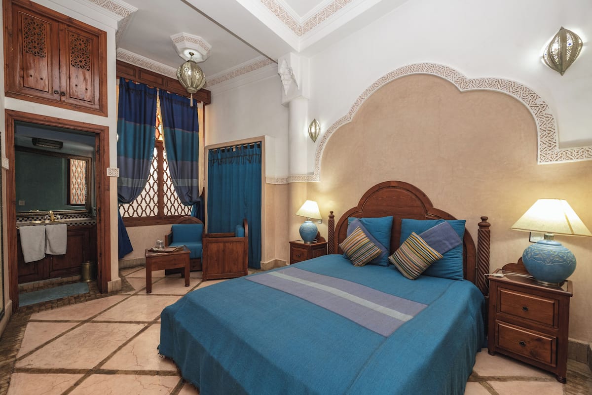 Une chambre avec un grand lit, une table de chevet en bois, une lampe, une petite table, une chaise, une armoire et une fenêtre avec des rideaux.