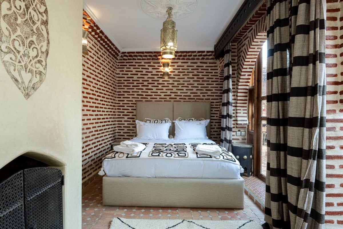 Une chambre d’hôtel avec un mur orné d’un motif en briques, un lit avec une literie blanche, un plaid à motifs et un luminaire suspendu.