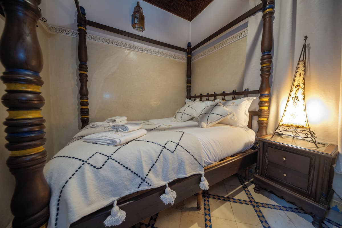 Un lit à baldaquin avec une couvre-lit à motifs noirs et blancs, une table de chevet en bois avec une lampe, et un sol carrelé avec un motif bleu et blanc.